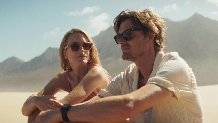 Islands: A White man and woman in sunglasses sit in a sunny desert landscape