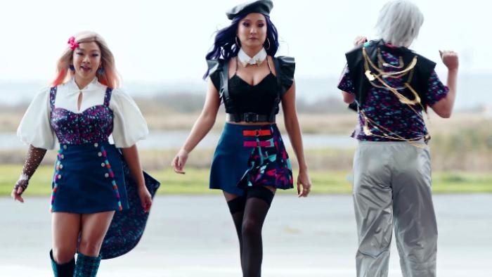 Joy Ride: Three Asian women are dressed in K-pop clothing and striding toward the camera on a tarmac. One wears a tilted hat, another has bleached-white hair, and the third has pink streaks and thigh-high boots. 