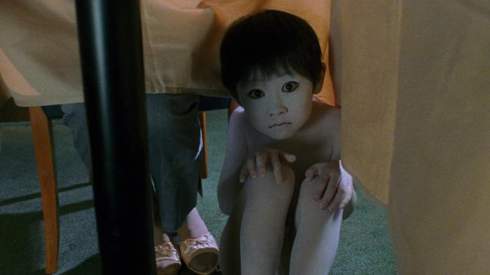 Ju-On—The Grudge: A ghostly child with wide eyes crouches under a table, looking forward