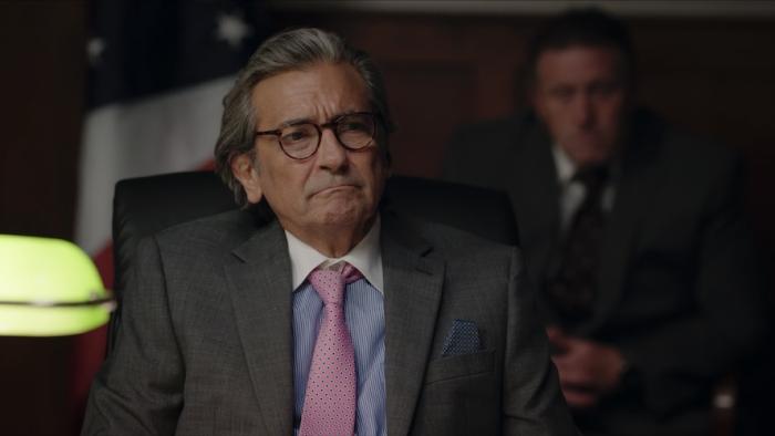 Junction Movie: Seated, wearing a suit and pink tie, Lawrence (Griffin Dunne) scrunches up his face in disdain