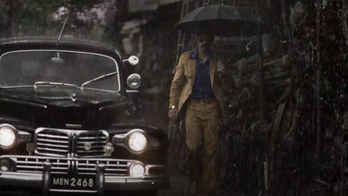 K.G.F -- Chapter 1: An Indian man in a yellow-brown suit holds an umbrella in the rain while next to a black car