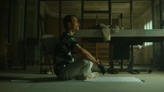 The Killer Movie: Seen from a side view, the Killer (Michael Fassbender) sits cross-legged on a white cloth, wearing his Hawaiian print shirt and black gloves, appearing to contemplate something