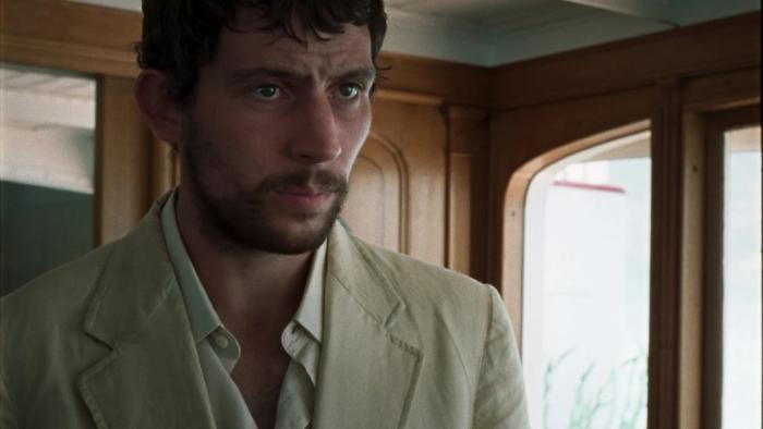 La Chimera: Josh O'Connor wears a light-colored linen suit as he stands in the wooden-clad cabin of a boat