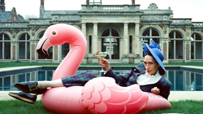 Lady: A White woman with dark brown hair poses on an inflatable flaming, holding a cocktail, in front of the swimming pool of a stately home