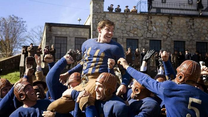 A rugby team holds up John Krasinski on their shoulders, cheering.