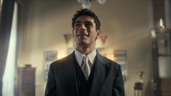 Lefter: The Story of Ordinarius movie: A Greek Turkish man with short black hair and in dark suit and tie, center, stands inside large office