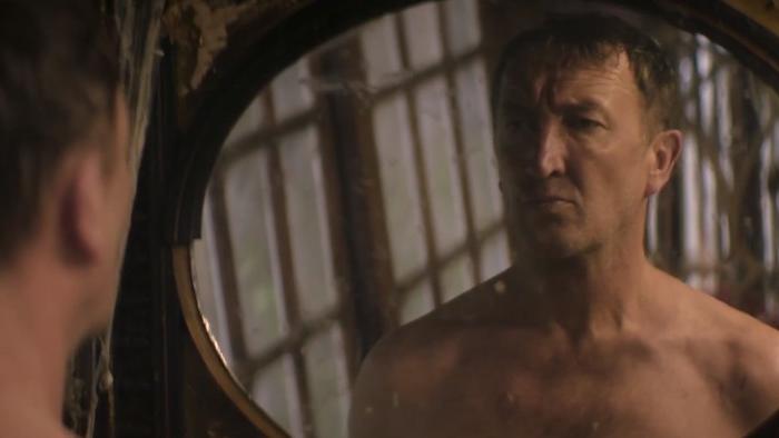 Lord of Misrule: A White middle-aged man stands shirtless in front of a mirror looking at his reflection.