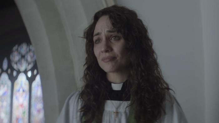 Lord of Misrule: A White female vicar stands in pulpit in vicars clothing looking upset.