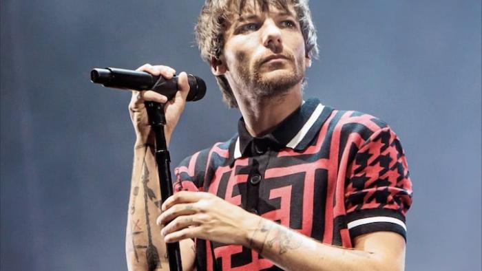 Louis Tomlinson -- All of Those Voices: The head and shoulders of Louis Tomlinson as he looks to his left, holding a microphone