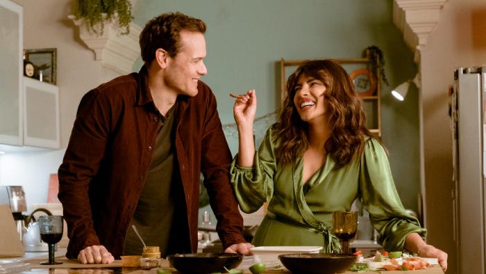 Love Again Movie: Priyanka Chopra Jonas, wearing a green wrap dress, laughs while offering a taste of the dish she's cooking to Sam Heughan