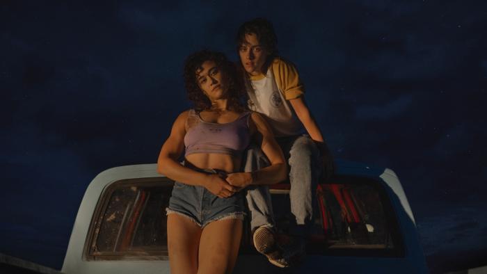 Love Lies Bleeding Movie: Katy O'Brian and Kristen Stewart sit close to each other on  the back of a truck