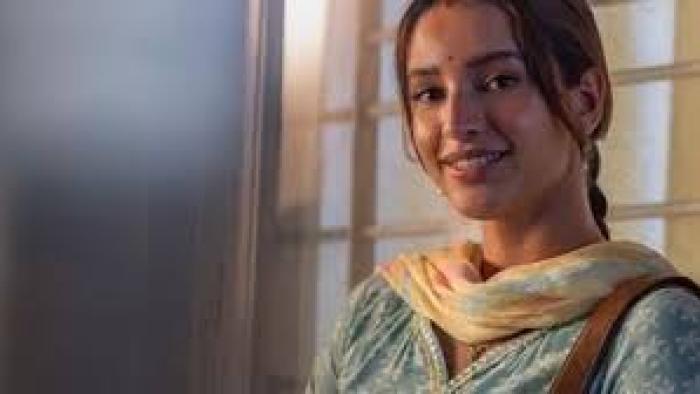 Maa Behen: Smiling Indian woman in traditional attire, standing near a window.