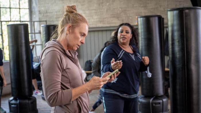 Mafia Mamma Movie: Toni Colette is in a gym looking with shock at her cell phone while her Black female friend is talking to her