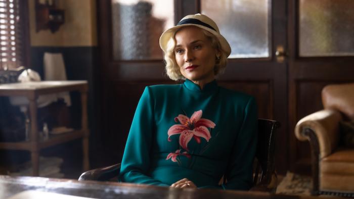 Marlowe Movie: Clare Cavendish (Diane Kruger) sits in a chair in Marlowe's office with a sly expression on her face; she wears a white hat and a blue dress with a pink flower on the front