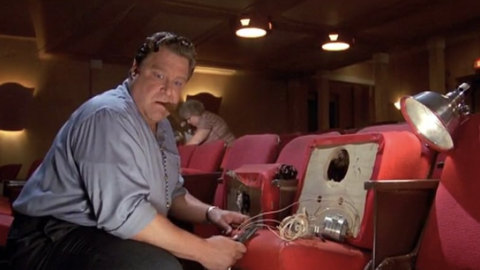 Matinee: Man sits in front of technical equipment with a cigar in his mouth on the red seats of a movie theater