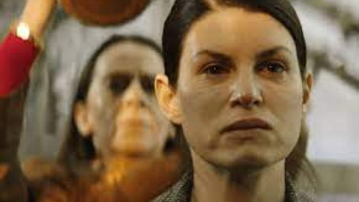 Close-up of a woman looking tense as another woman behind her raises an unknown object overhead.