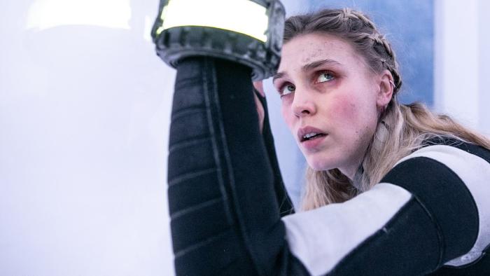 Meander: A young White blond woman holds up an illuminated bracelet, crouched in a small, bright white space.