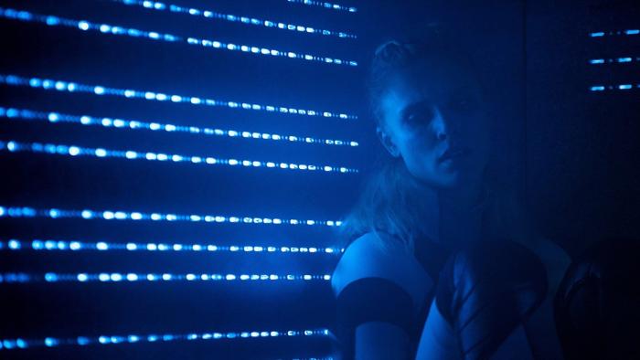 Meander: A young White blond woman leans back against a wall, with blue light shining through small slats in the darkness.