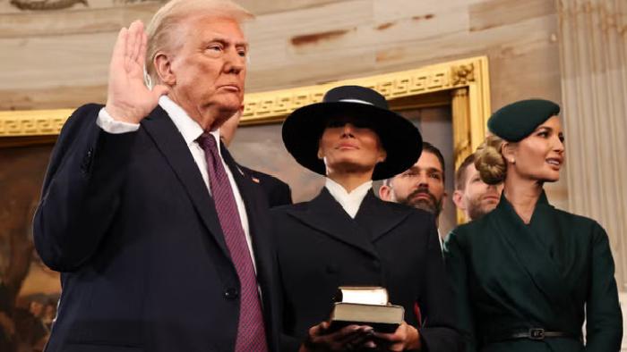 Melania movie: President Trump holds up his hand at his swearing-in ceremony; Melania holds his hand and obscures her eyes with a hat