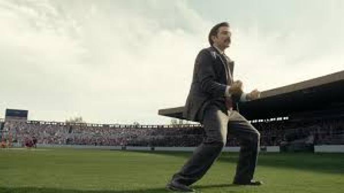 Mexico 86 Movie: Diego Luna in suit energetically cheering on a soccer field with a crowded stadium.