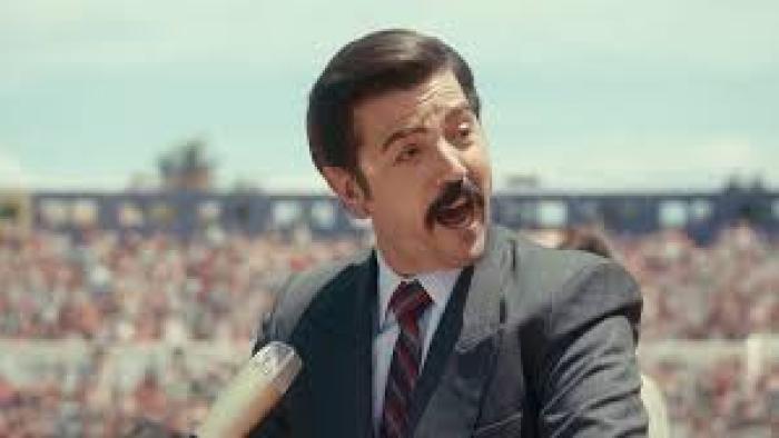 Mexico 86 Movie: Diego Luna in a suit speaking into a microphone at an outdoor event.