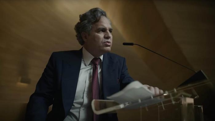 Mickey 17 Movie: In a suit and red tie, religious leader Kenneth Marshall (Mark Ruffalo) gives a speech