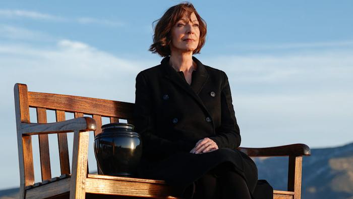 Miss You, Love You: Allison Janney sits on a bench outside in a black coat, next to an urn