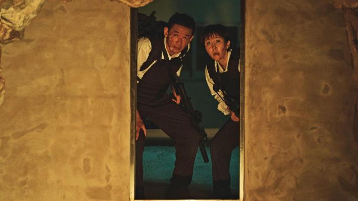 Mission: Cross movie: Korean man, woman, both with guns and in riot gear, center, peer through darkly lit doorway