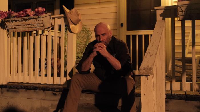 Mob Land Movie: Sheriff Bodie Davis (John Travolta) sits on a front porch at night, his hands folded and held up to his face, looking concerned