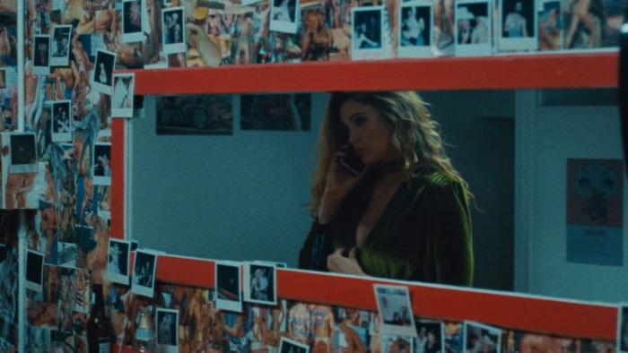 Monica Movie: A woman on the phone through the reflection in a rectangular mirror hanging on a wall full of Polaroid pictures