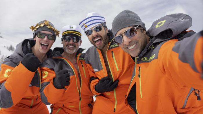 Mountainhead: Four men in matching orange snowsuits, grinning and posing for a selfie on a snowy mountain