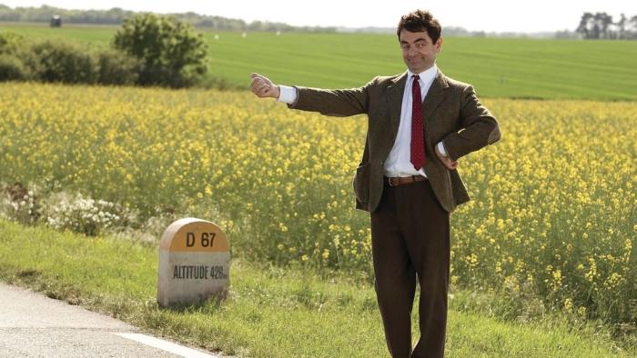 Rowan Atkinson as Mr. Bean stands on the side of the road in a suit, thumb up. There is a small stone next to him that reads "D 67, Altitude 466"