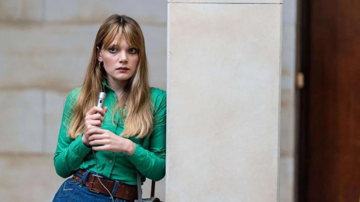 Mr. Car and the Knights Templar movie: A female reporter holding a microphone leans against a wall while wearing a green shirt. She has a serious look on her face