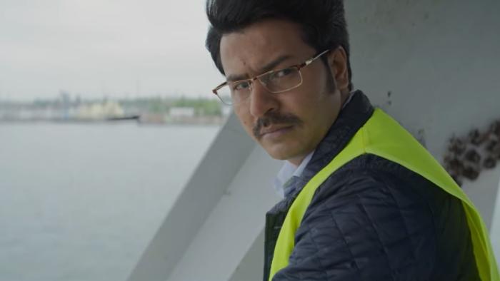 Mrs. Chatterjee vs. Norway: A middle-aged Indian man looks over shoulder wearing yellow hi-vis vest with water and steel girder in background