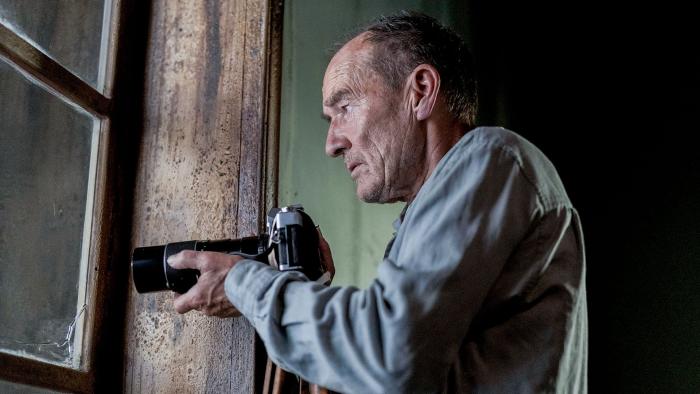 My Neighbor, Adolf: An elder White man stands in front of a window, holding a camera