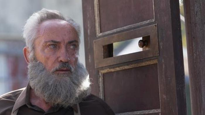 My Neighbor, Adolf: A White bearded-man stands next to a wooden door, his eyes in line with a peephole