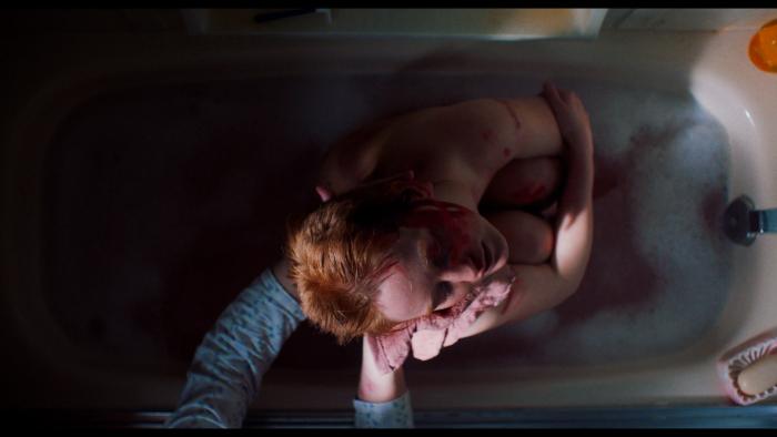 My Animal Movie: A young redheaded woman clutches her knees tightly in a bathtub while a woman's hands are seen, washing her cuts and bruises