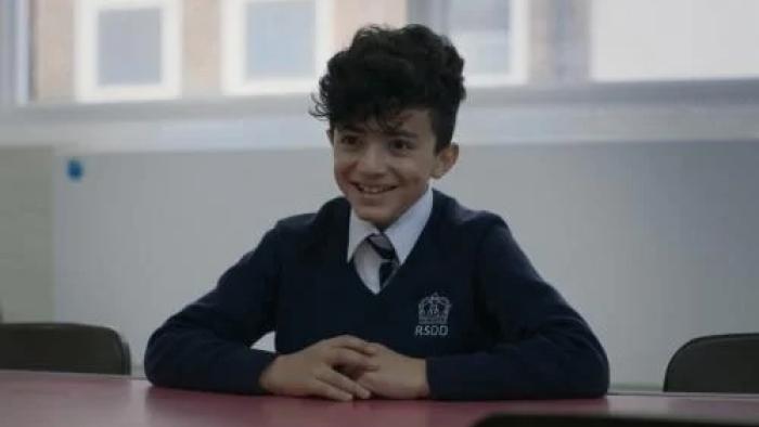 Name Me Lawand: A young boy smiles as he sits at a desk wearing a school uniform