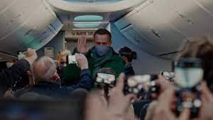Navalny: A Russian white man center walks down airplane aisle while other passengers all face him with their camera phones filming him