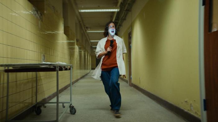 Nightwatch: Demons Are Forever movie: Danish white woman in white lab coat, headphones, and flashlight sings in hallway
