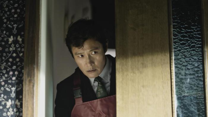 No Other Choice Movie: Wearing burgundy-colored waders, Man-su peeks through a doorway, looking wary