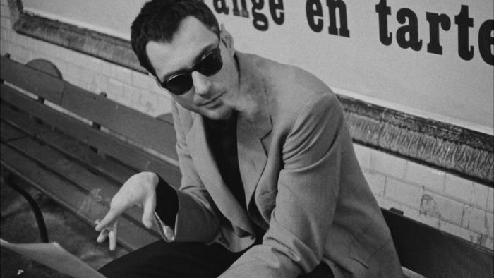 Nouvelle Vague Movie: In dark glasses, Godard sits on a subway bench, smoking and working on pages for his movie