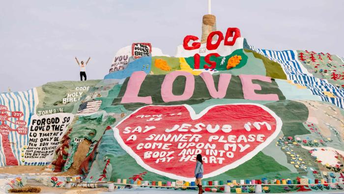 Off the Record movie: Ryan Hansen stands atop an art display on a hill sayihg "God Is Love"; Rainey Qualley looks up at him