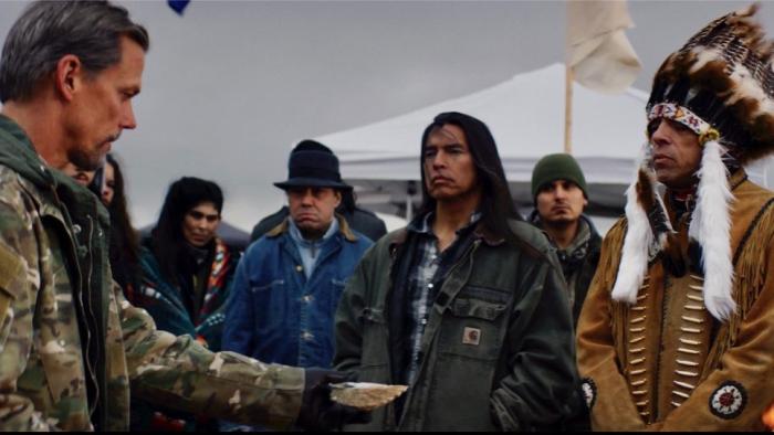 On Sacred Ground Movie: A White man in Army fatigues holds out an offering to a First Nation chief and other Native American men