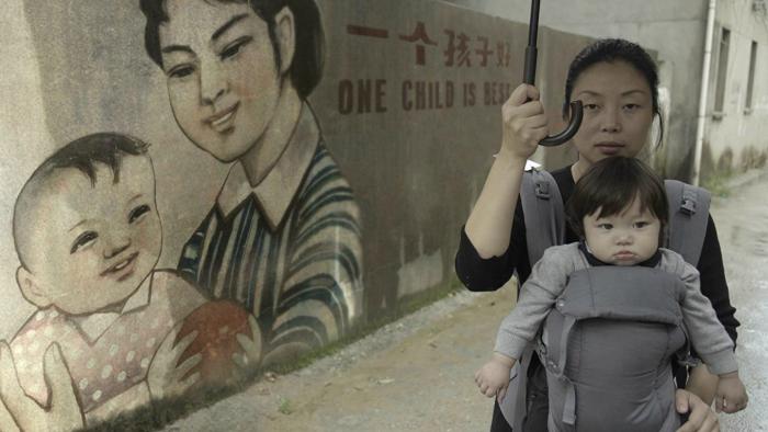 One Child Nation Movie: A mother holds her baby in a carrier in front of a mural that reads "One child is best"