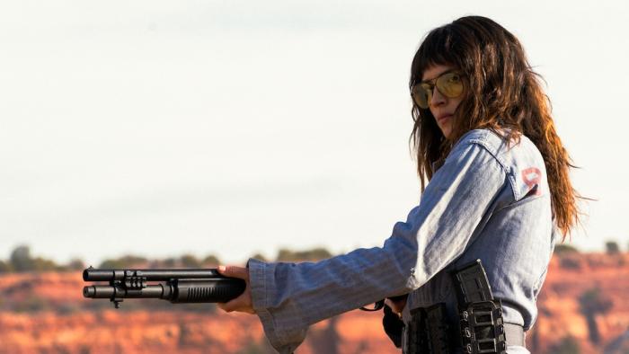 Onslaught Movie: Wearing shaded glasses, Celeste holds a high-powered rifle as she shoots targets in the desert