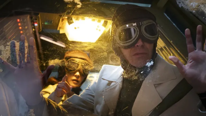 Our Times movie: Mexican woman background, man foreground, both wearing aeronautical headgear in time capsule machine, yellow light above them