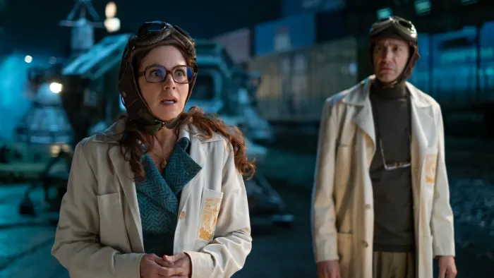 Our Times movie: Mexican woman left, man right, both in lab coats and aeronautical headgear stand in front of large time capsule machine at night