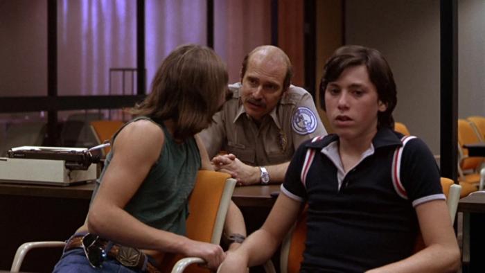 Over the Edge: A young man turns to talk to a police officer as his friend sits beside him in a police station