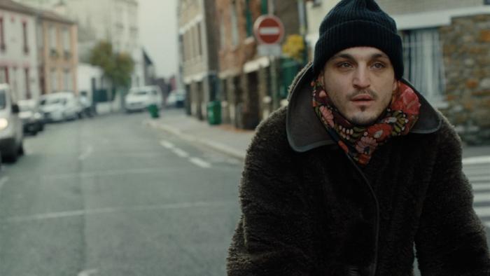 Passages: A young White man wearing a dark hat and coat as he rides a bicycle through the streets.